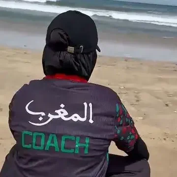 qui-est-coach-muslima-fit Coach sportive pour femmes Muslima Fit assise sur la plage, portant un vêtement avec écrit "المغرب" et "COACH" au dos.